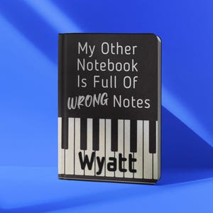 May include: A black notebook featuring a piano keyboard graphic at the bottom and the white text My Other Notebook Is Full Of WRONG Notes with the name Wyatt printed in black on the keys. The cover has a matte finish and a vertical spine.