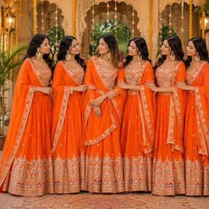 May include: Five matching bright orange lehengas featuring intricate gold embroidery and mirror work along the hemline. Each set includes a sleeveless blouse, a floor-length pleated skirt, and a coordinating sheer dupatta draped over the shoulder.
