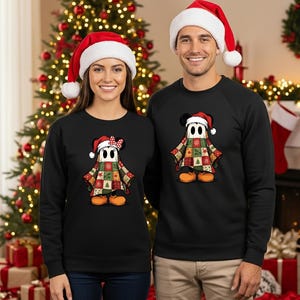 May include: Two black crewneck sweatshirts featuring a graphic of a ghost character wearing a Santa hat and covered in a colorful patchwork quilt with holiday patterns like stars, trees, and holly. The ghost has orange shoes and mouse ears.