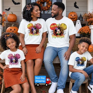 May include: A group of four people wearing white t-shirts with Disney character Halloween graphics. The shirts feature Minnie, Mickey, Daisy, and Donald Duck in witch hats with pumpkins and trees. The group wears skirts and jeans.