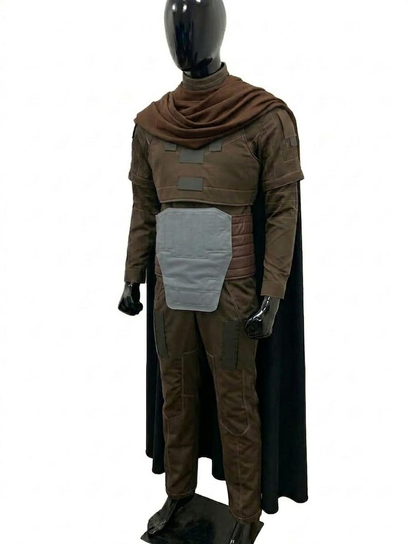 May include: A brown long-sleeved costume set featuring a dark brown cowl, a chest plate, a light grey abdominal armour piece, and a long black cape. The outfit includes matching brown trousers with reinforced panels and is displayed on a black mannequin.