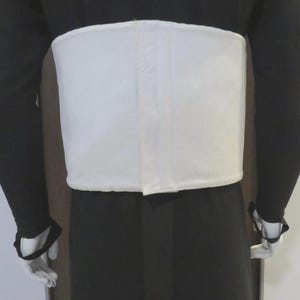 May include: A wide, white, rectangular abdominal support belt worn over a black long-sleeved garment. The belt features a central vertical seam and a smooth, solid white fabric construction that wraps around the midsection of the torso.