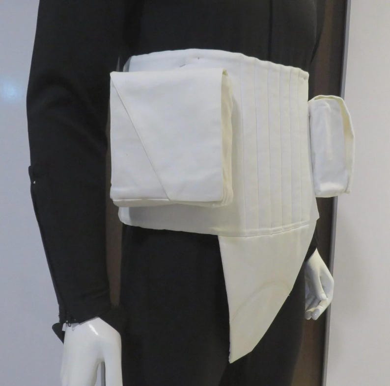 May include: A white utility belt featuring a wide, structured waistband with vertical stitching, a large square pouch on the front, and a smaller rectangular pouch on the side. The belt includes a pointed fabric panel extending downward from the center.