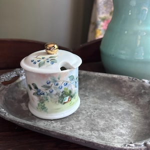May include: A white ceramic sugar bowl with a lid, featuring a hand-painted design of blueberries and a small house in a landscape. The lid has a gold knob and a slot for a spoon. The bowl sits on a silver tray.