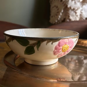 May include: A small, off-white ceramic bowl with a floral design. The bowl features a pink flower with a yellow center and green leaves. The rim is outlined in a dark brown color.