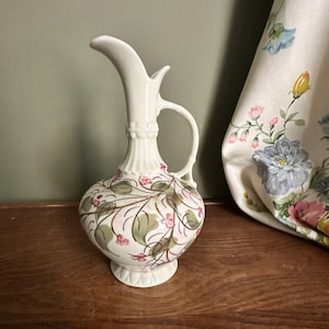 May include: A cream-colored ceramic pitcher with a slender, curved neck and a single handle. The rounded body features a hand-painted pattern of green leaves, thin brown vines, and small pink flowers. The base and neck have a fluted design.