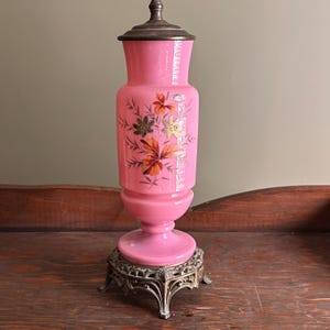 May include: A pink glass vase with a silver-colored lid and base. The vase features a floral design in shades of orange, gold, and brown. The vase is approximately 12 inches tall and sits on an ornate metal base.