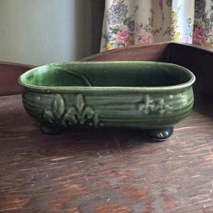 May include: A green ceramic planter with a rectangular shape and three small feet. The planter features embossed fleur-de-lis designs and horizontal ridges. The interior is smooth, and the exterior has a glossy finish.