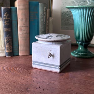 May include: A square, ceramic container with a lid, featuring a light gray base with blue-gray stripes. The lid has a decorative pattern. A small, metal pull is on the front. Books and a green vase are in the background.