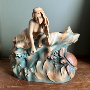 May include: A ceramic figurine featuring a reclining figure with long hair resting on a stylized wave formation. The piece is finished in muted tones of beige and teal, with a small sea turtle sculpted into the lower right side of the base.