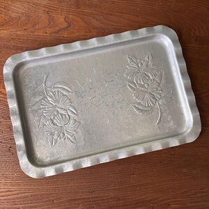 May include: A rectangular, silver-colored metal tray with a scalloped edge. The tray features embossed floral designs on the surface. The tray is set on a wooden surface.