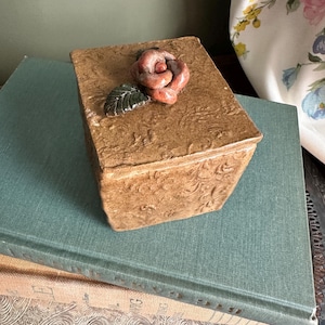 May include: A square ceramic box with a textured tan surface featuring an embossed floral pattern. The lid is adorned with a sculpted, glossy reddish-brown rose and a single dark green leaf, providing a decorative accent to the container.