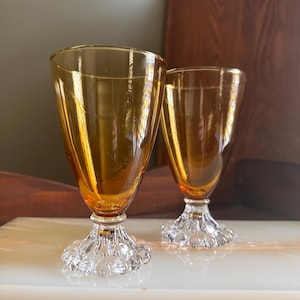 May include: Two amber-colored vintage wine glasses. The glasses have a conical shape and sit on a clear, bubble-like base. The stem is short and the glass has a smooth, reflective surface. The glasses are displayed on a white surface.