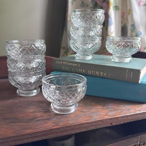 May include: A collection of clear, diamond-cut glass bowls. Some are stacked, others are arranged on a wooden surface and a book. The bowls have a pedestal base and a decorative, textured pattern. The book's title is "Jack O'Connor's Big Game Hunts".