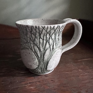 May include: A speckled ceramic mug with a detailed tree design carved into the surface. The mug has a large handle and a wide opening. The color is a speckled off-white with a gray tree design. The mug is sitting on a wooden surface.
