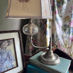 May include: A vintage table lamp with a floral patterned lampshade and a clear glass globe. The lamp sits on a metal base, next to a framed portrait and two books. The books are teal and the portrait is in a dark wooden frame.