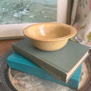 May include: A small, round, yellow ceramic bowl with a speckled finish and a wide, flat rim. The bowl features a smooth, glossy glaze with dark brown flecks distributed across the surface, creating a textured appearance on the exterior and interior.