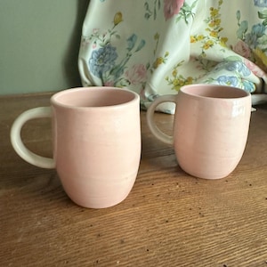 May include: Two matching pale pink ceramic mugs with a smooth, matte finish and rounded bodies. Each mug features a simple, curved handle extending from the side. The mugs have a consistent, soft pastel color throughout their exterior surfaces.