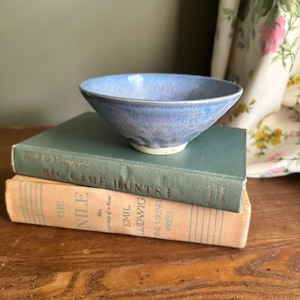 May include: A small, round, light blue ceramic bowl with a smooth, glossy finish and a white base. The bowl features subtle variations in color, ranging from pale sky blue to deeper periwinkle tones, and sits atop two stacked hardcover books.