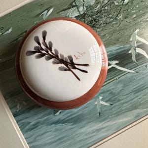 May include: A round, ceramic trinket box with a white lid and a reddish-brown base. The lid features a watercolor painting of a sprig of pussy willow branches in shades of gray and brown, with the word "kehu" in red.