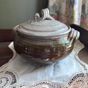 May include: A handcrafted ceramic lidded serving bowl. The bowl is glazed in shades of brown and white, with a textured lid and decorative handles. The bowl is sitting on a white lace doily.