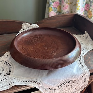 May include: A circular wooden bowl with a deep brown finish and a prominent, swirling natural wood grain pattern. The bowl features a smooth, rounded rim and a shallow, concave interior surface, showcasing the organic texture of the wood.