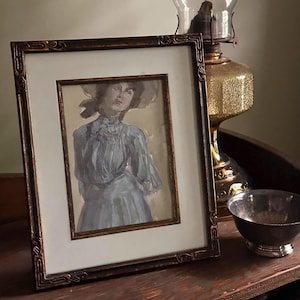 May include: A framed watercolor painting of a woman in a blue dress and hat. The painting is in a dark brown ornate frame. A vintage oil lamp and a silver bowl are next to the painting.