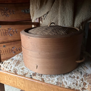 May include: A round, light brown bamboo steamer with a woven lid and a small handle. The steamer has a cylindrical shape and is sitting on a patterned chair.
