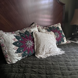 May include: A bed with a dark brown headboard and a dark green quilted bedspread. Three decorative pillows are arranged on the bed. Two pillows have a floral design with burgundy and green accents. A white crocheted pillow with fringe is in the center.