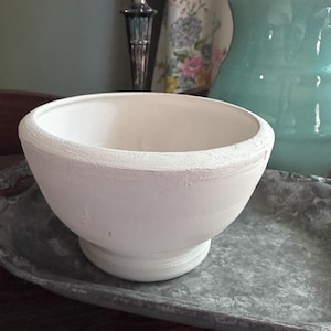 May include: A white ceramic bowl with a rounded shape and a pedestal base. The bowl has a textured rim and sits on a gray metal tray. The background includes a green pitcher and a floral patterned fabric.