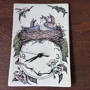 May include: Rectangular ceramic clock with a painted design of a bird's nest with three birds and a clock face. The clock hands point to 9:15. Floral and leaf accents surround the clock face. The clock is on a wooden surface.