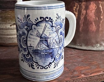 May include: A white ceramic mug with a blue windmill and floral design. The mug has a handle and is decorated with a traditional Dutch Delftware pattern. The mug is cylindrical and has a slightly flared rim.