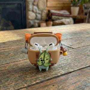 Puede incluir: Una funda marrón para AirPods con un diseño de dibujos animados. La funda está abierta, revelando auriculares blancos en el interior. La funda tiene un diseño de hoja y hongo. La funda está sobre una mesa de madera.