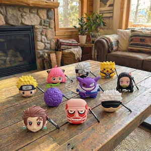 May include: A collection of character-themed AirPod cases in various colors, including yellow, pink, purple, and brown. The cases feature designs from popular anime and video games, sitting on a rustic wooden table.