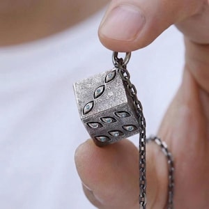 May include: A silver-colored pendant necklace featuring a cube-shaped charm. The charm is textured and has eye-shaped designs with blue accents on each face. The necklace chain is also silver-colored.