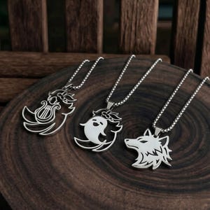May include: Three silver-colored pendant necklaces with different designs: a lyre, a ghost, and a wolf head. Each pendant hangs from a delicate chain. The necklaces are displayed on a dark wooden surface.