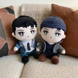 May include: Two plush dolls of a male character with black hair and a serious expression. One doll wears a blue jacket, white shirt, and black tie. The other doll wears a blue beanie and a gray jacket. Both dolls are sitting on a beige couch.