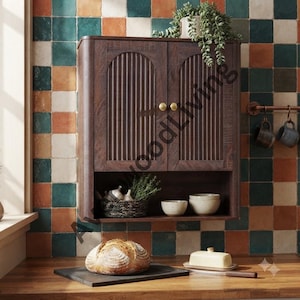 May include: A dark brown wooden wall cabinet with two arched doors featuring vertical fluted panels and small round gold knobs. The cabinet includes an open lower shelf containing two ceramic bowls and a woven basket with greenery.