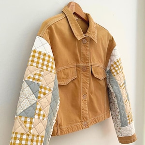 May include: A tan denim jacket featuring long quilted sleeves with a patchwork design. The sleeves incorporate a mix of yellow and white gingham, solid sage green, and cream floral fabric patterns. The jacket has a collar and button front.