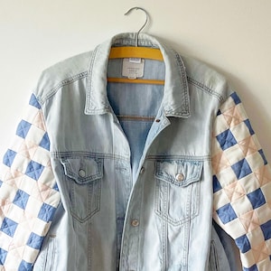 May include: A light-wash blue denim jacket featuring long sleeves with a quilted, checkered pattern in shades of light blue and soft pink. The jacket includes a classic collar, two button-flap chest pockets, and a front button closure with an American Eagle label.