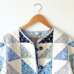 Könnte beinhalten: Eine gesteppte Jacke mit einem Patchwork-Design aus cremefarbenem Stoff und verschiedenen blauen, weißen und grünen Blumenmustern. Das Kleidungsstück hat einen runden Ausschnitt, eine Knopfleiste vorne mit dunklen Knöpfen und ein blau-weiß gestreiftes Futter.