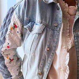 Può includere: Una giacca in denim dal lavaggio chiaro con maniche lunghe caratterizzate da pannelli bianchi trapuntati e sezioni in velluto a coste blu. Il tessuto bianco trapuntato è decorato con ricami floreali colorati. Giacca con colletto e bottoni.
