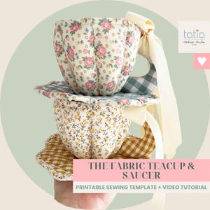 Peut inclure: Deux tasses et soucoupes en tissu empilées avec des motifs floraux et des accents vichy. La tasse supérieure présente des roses roses sur fond crème, tandis que la tasse inférieure a des fleurs jaunes. Elles incluent un ruban crème et le texte THE FABRIC TEACUP & SAUCER.