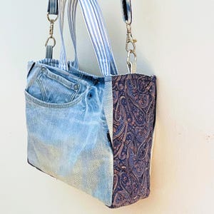May include: A tote bag constructed from light blue denim fabric featuring a visible pocket and metal rivets. The side panel is made of dark blue fabric with a detailed paisley pattern. It includes light blue and white striped fabric handles.