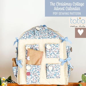 Peut inclure: Calendrier de l'Avent en tissu en forme de maison, avec des poches à imprimé floral et des nœuds bleus. Le texte "The Christmas Cottage Advent Calendar PDF Sewing Pattern" est affiché sur fond bleu. Le calendrier a des boutons en bois et contient de petits paquets.