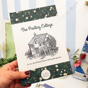 May include: A journal titled "The Posting Cottage" with a floral pattern on the top and bottom edges. The cover displays a black and white cottage illustration. The journal is held in a hand.