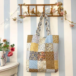 Op de afbeelding: Een patchwork tote bag met een verscheidenheid aan bloemen- en patroonstoffen vierkanten. De tas heeft gevlochten handvatten en hangt aan een houten rek. De achtergrond is een gestreepte muur met een bloemenslinger.