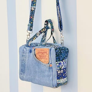 Puede incluir: Bolso cruzado de mezclilla azul claro, con una etiqueta Levi Strauss & Co. y un bolsillo delantero. El bolso tiene detalles de tela estampada floral y una correa ajustable a juego. El bolso cuelga contra una pared a rayas azules y blancas.