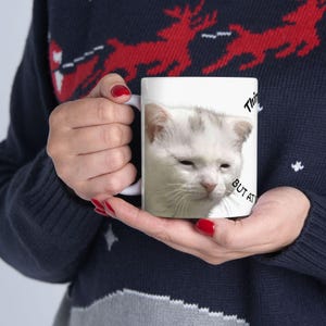 May include: White ceramic mug featuring a close-up of a sleepy-looking white and gray cat. The mug has the text "THINK BUT AT" printed on it. The mug is held by a person wearing a navy blue sweater with a red reindeer pattern.