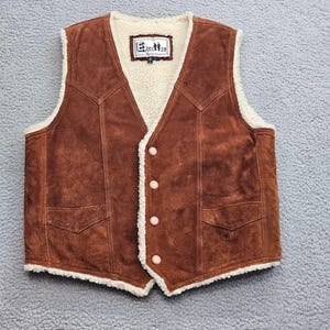 May include: A brown suede vest featuring a cream-colored faux shearling lining, a V-neckline, four front buttons, and two lower pockets. The interior label displays the text Excelled. The vest has a textured finish and visible stitching.